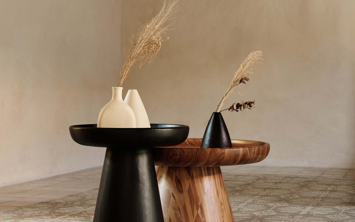 Two decorative side tables in a styled interior setting, one matte black and one natural wood, each holding a small vase with dried grass stems against a warm beige wall.