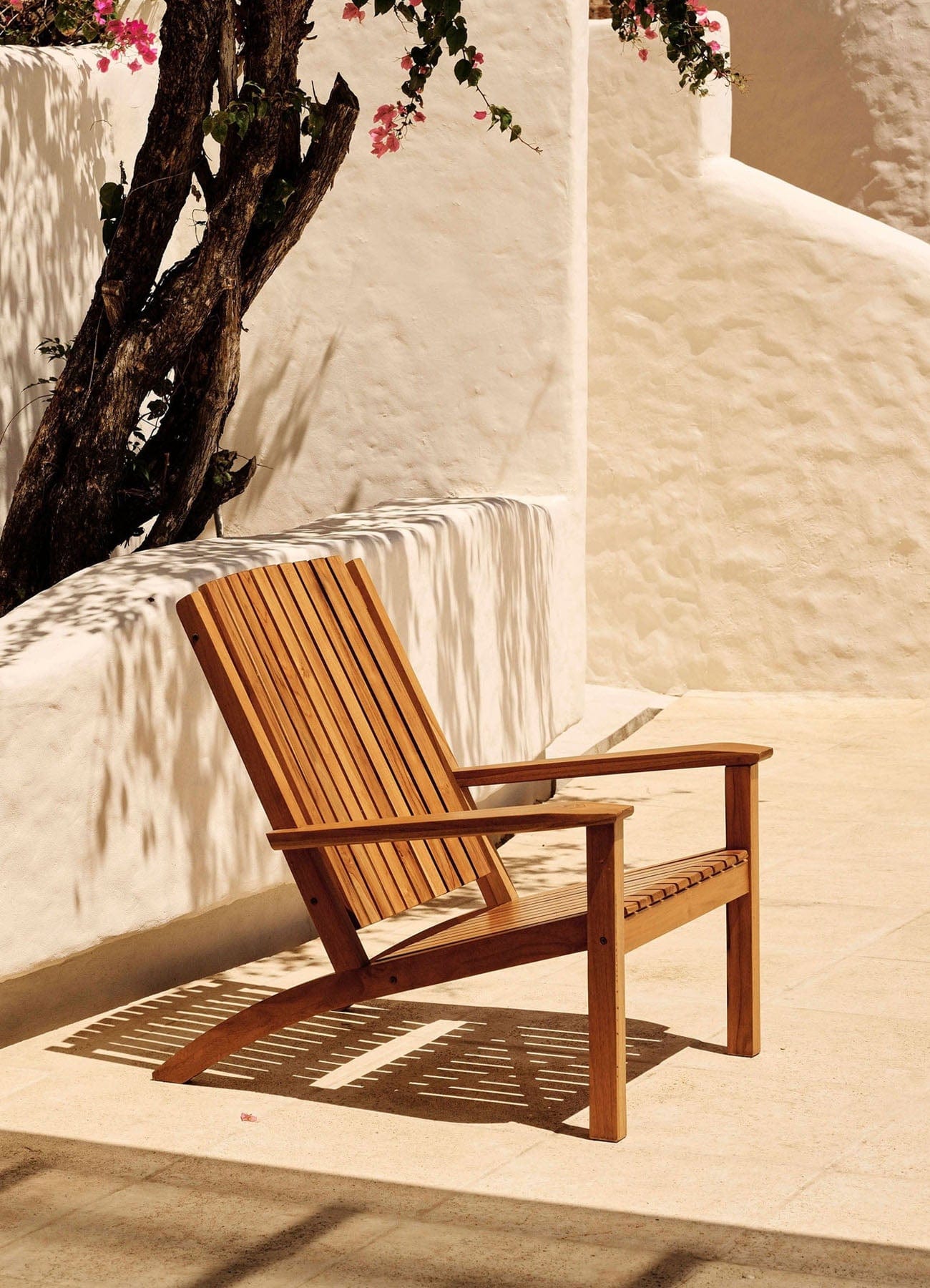 Wooden teak adirondack chair on a sunlit patio with white walls and a tree in the background