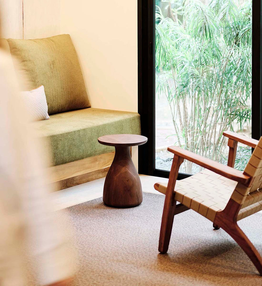 A styled interior corner featuring a low upholstered bench with a large olive-green cushion, a small round wooden side table, and a woven-back wooden chair with a warm wood frame, set on a textured rug beside a large window overlooking bamboo.