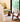 A styled interior corner featuring a low upholstered bench with a large olive-green cushion, a small round wooden side table, and a woven-back wooden chair with a warm wood frame, set on a textured rug beside a large window overlooking bamboo.