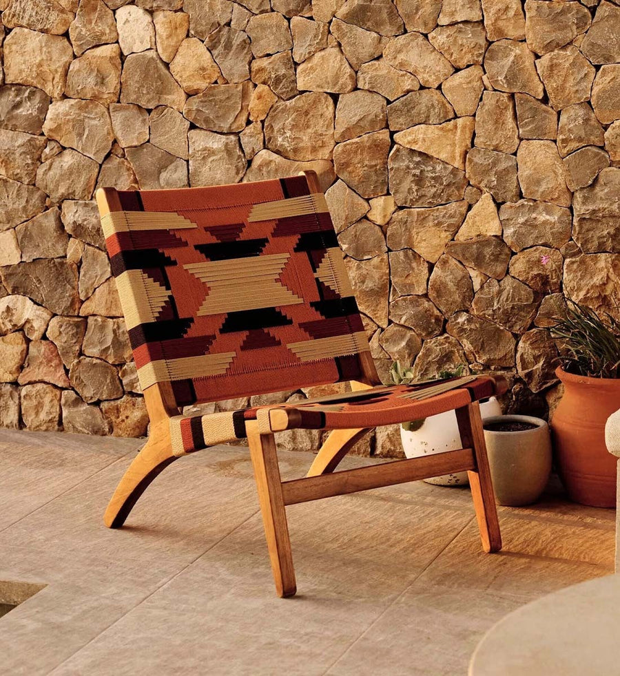 A mid-century modern wooden lounge chair with a woven geometric patterned seat and back, set against a rustic stone wall background.