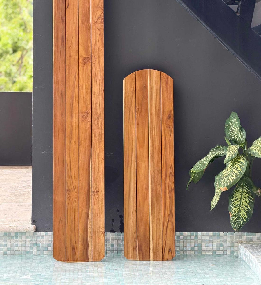 Two vertical wooden boards with rounded tops, showing natural wood grain, standing on a tiled surface against a dark wall with a green plant nearby.