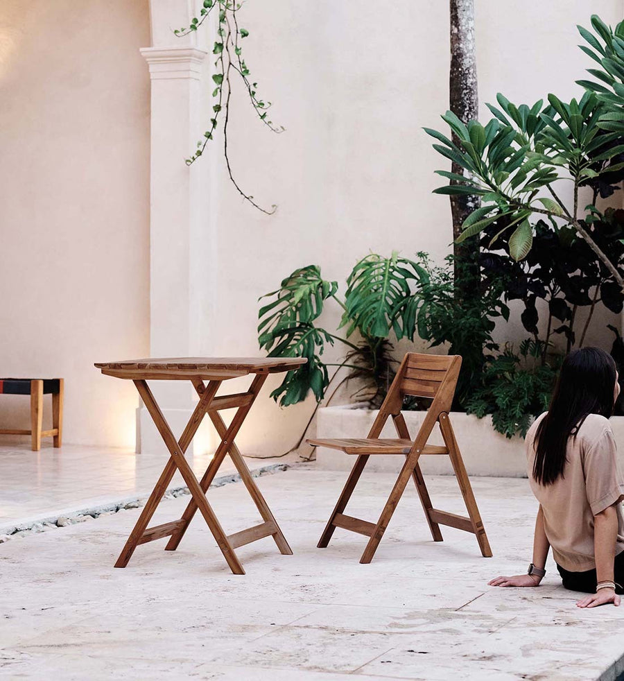 A wooden folding table and matching folding chair in a light brown finish, set on a stone patio with potted green plants and a woman sitting nearby.