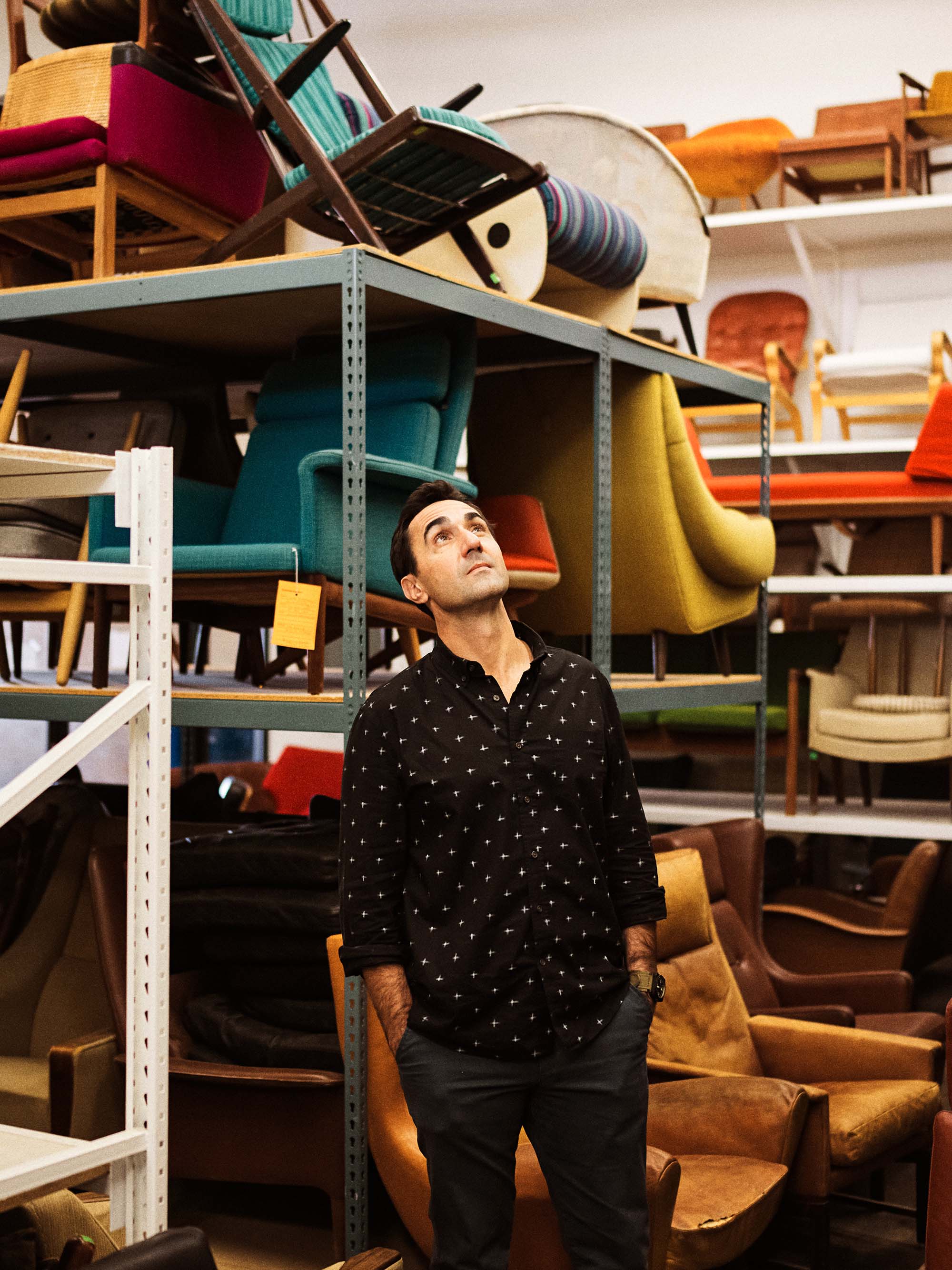 A man in a black patterned button-down shirt and dark pants stands in a furniture showroom or warehouse filled with shelving and colorful chairs and stools.