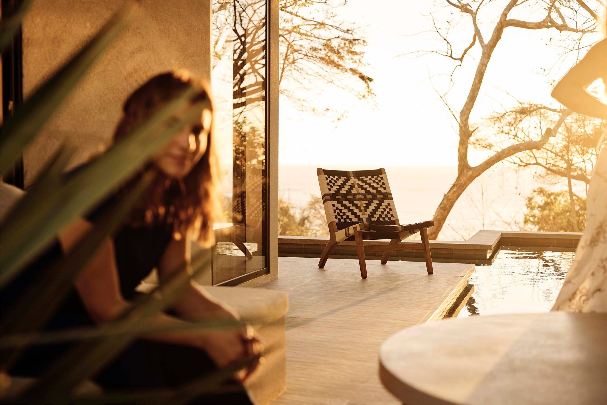 Woman sitting on a wooden deck with a view of a pool and trees during sunset.