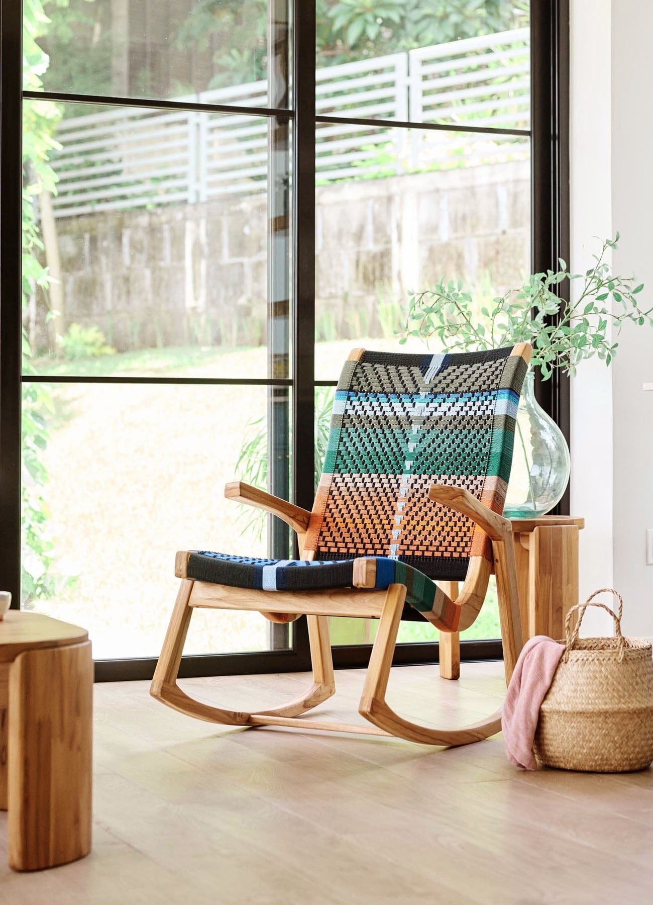 Wooden teak rocking chair with woven backrest in a room with large windows and plants.