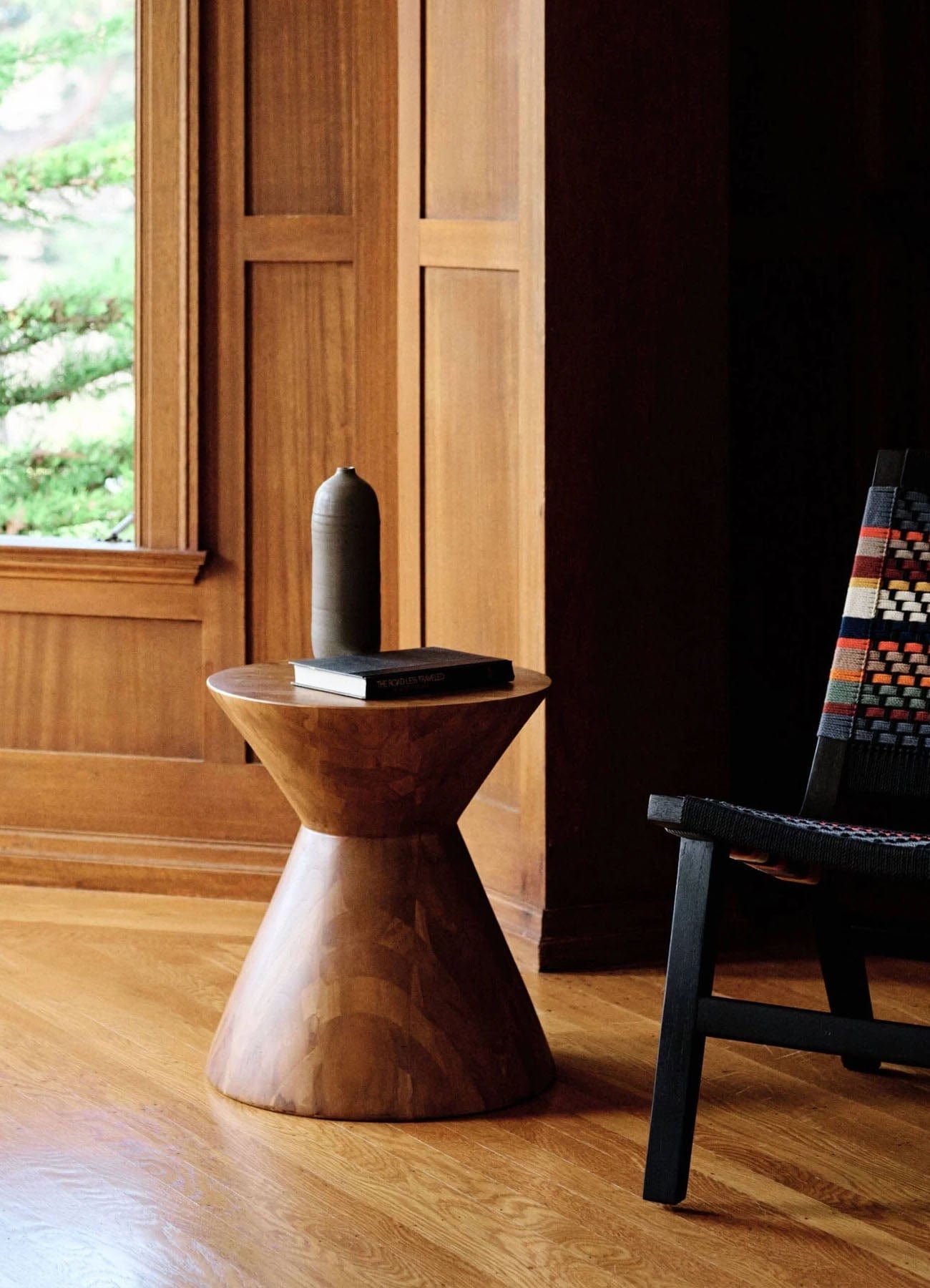 Wooden teak accent table/stool with a black vase on top in a room with wooden walls and a chair.