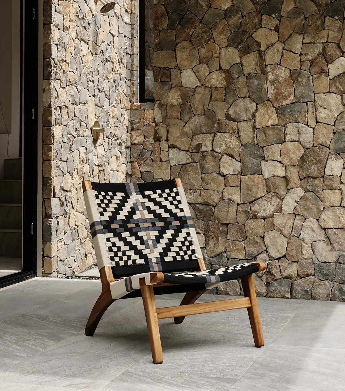 Wooden teak lounge chair with a patterned woven seat against a stone wall.
